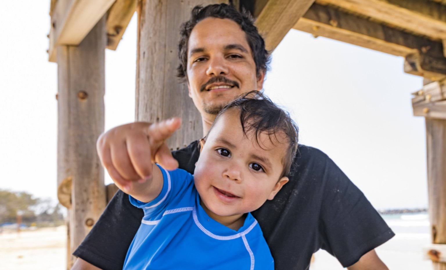 A father stands smiling at camera, his son is directly in front of him, smiling and pointing at the camera
