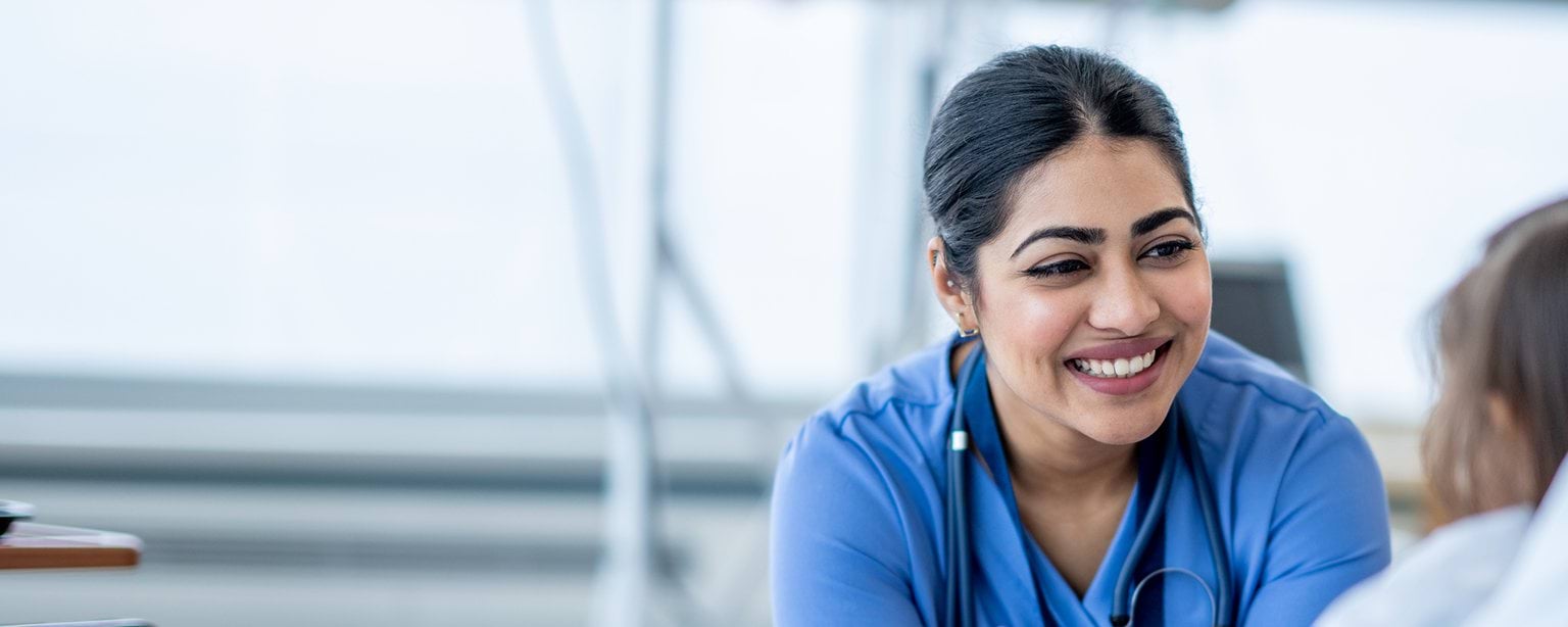 A nurse in blue scrubs and a stethoscope smiles while interacting with a patient.