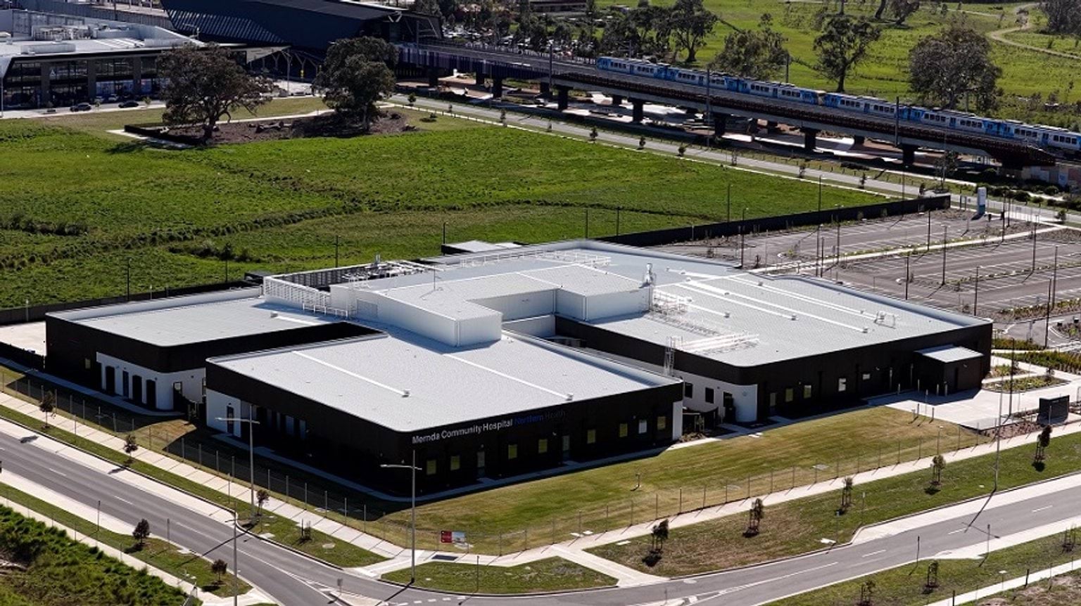 An aerial image of the Mernda Community Hospital building. It has a white tin roof and is surrounded by green grass.