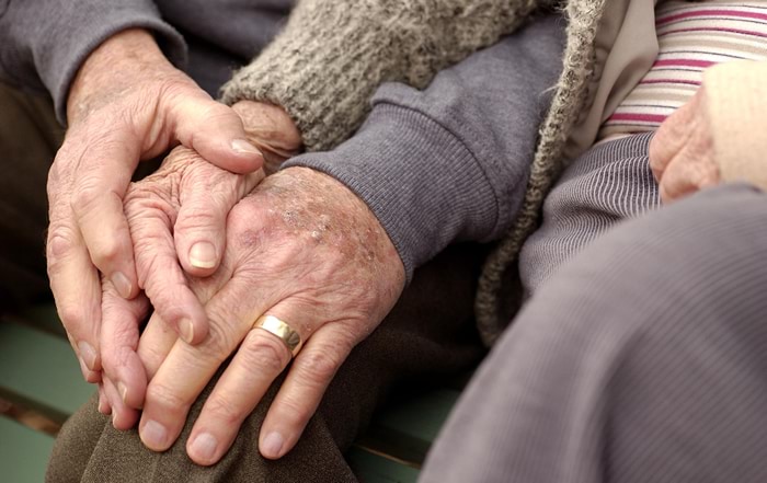 The photo is of hands of two older people, male and female, resting on the knee of the man. It demonstrates intimacy and comfort between the two people.